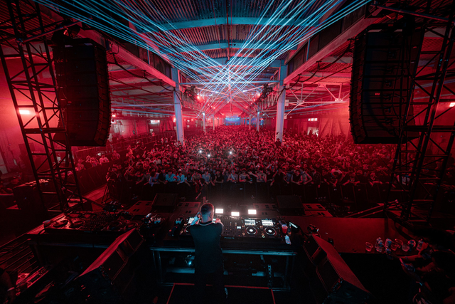 A DJ performs on stage facing a large crowd in a dark, industrial venue lit with red lights and blue laser beams overhead.